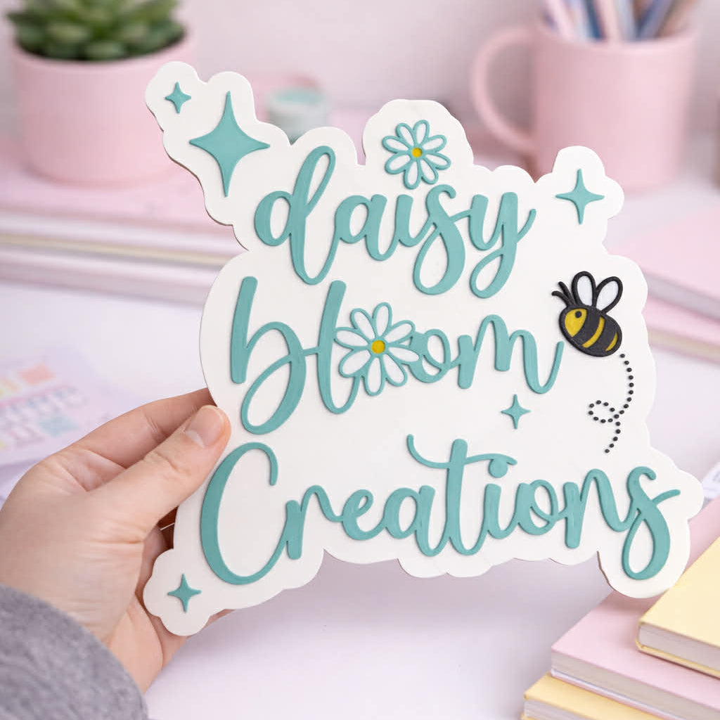 Hand holding a 'daisy bloom Creations' sticker with a desk and stationery in the background