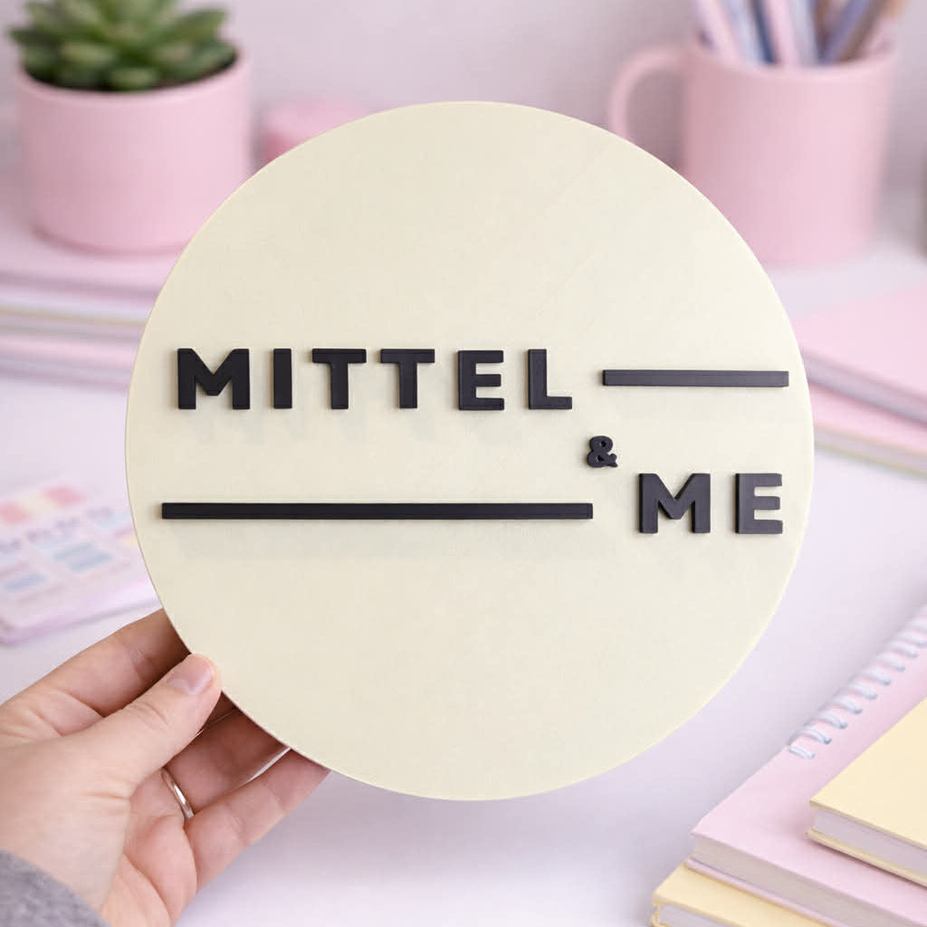 Round sign with 'MITTEL & ME' text held by a hand on a desk with stationery items.