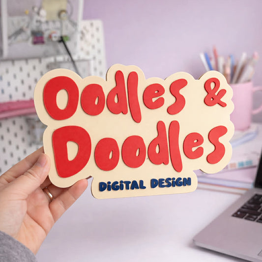 Hand holding a sign with 'Oodles & Doodles Digital Design' over a desk with stationery items.