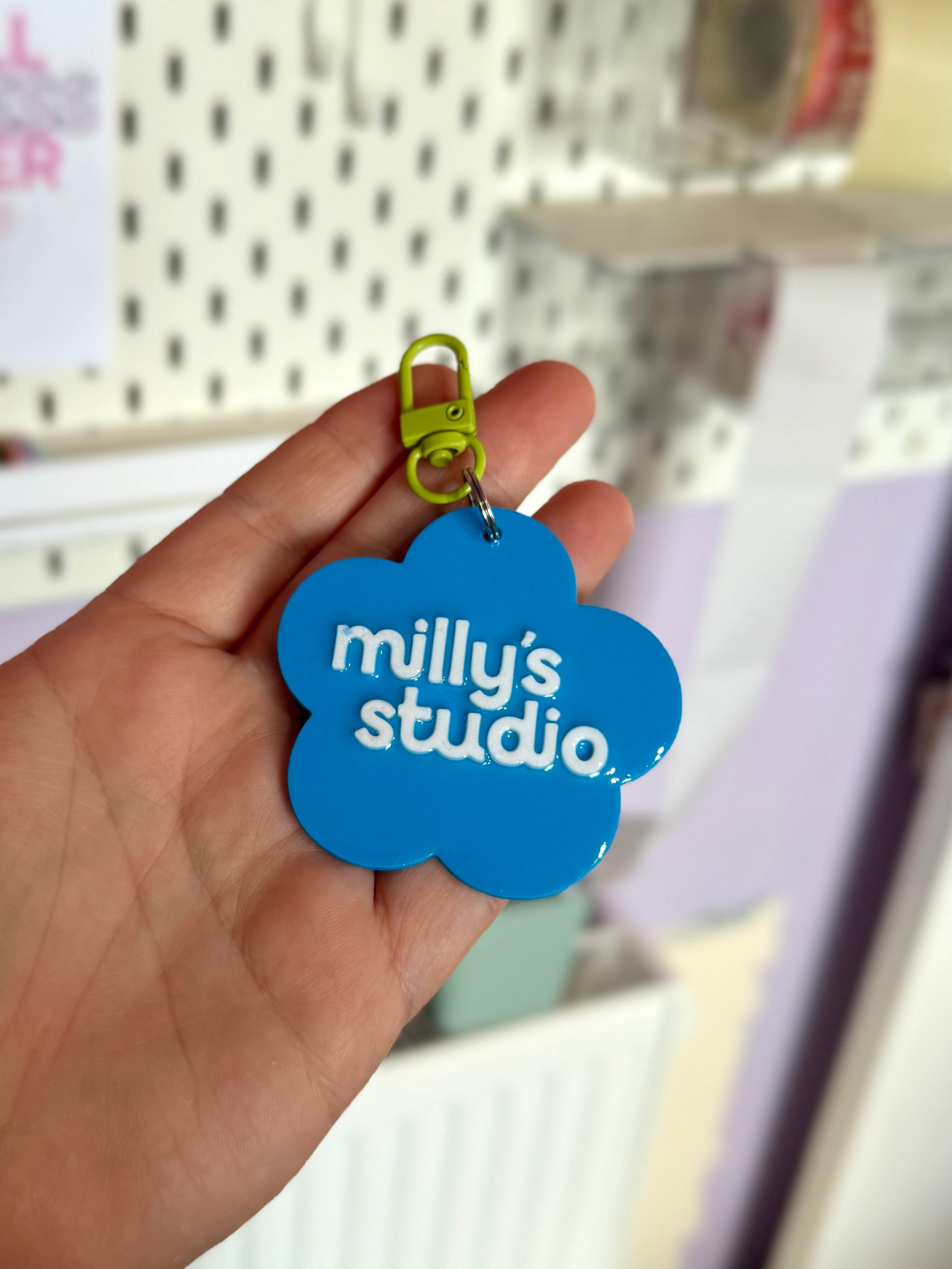 Blue cloud-shaped keychain with 'milly's studio' text held by a hand against a blurred background.