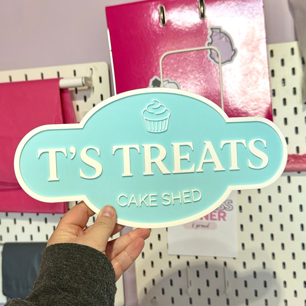Hand holding a sign that reads 'T's Treats Cake Shed' against a colorful background.